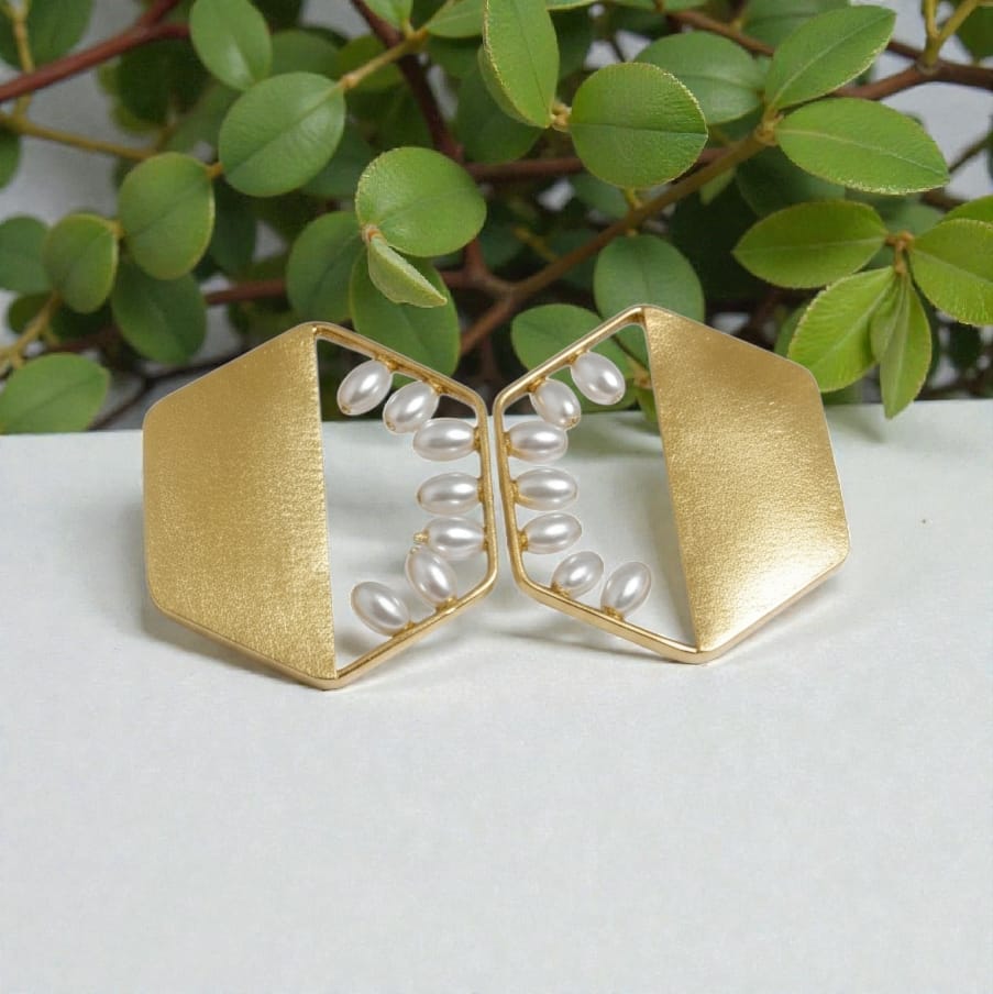 A pair of gold geometric cuff-style earrings with white faux pearls arranged in vertical strands, displayed on a light surface with green leaves in the background.