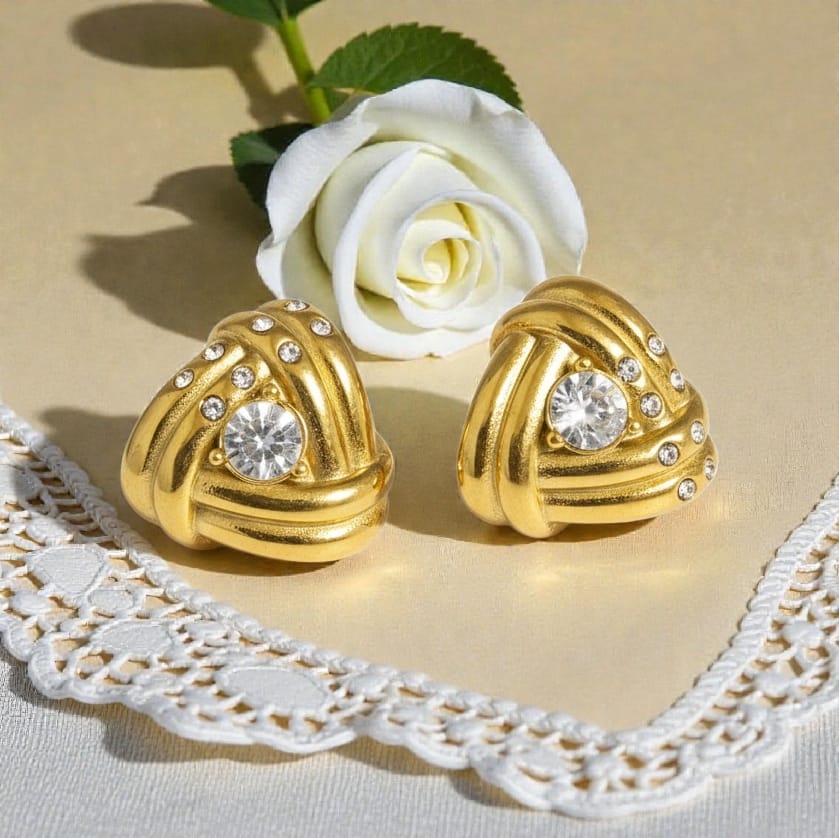 A pair of gold-toned heart-shaped earrings with clear gemstones, styled on a beige surface with a white rose and lace runner.