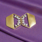 A pair of gold geometric earrings with white faux pearl accents, displayed on a purple satin fabric background.