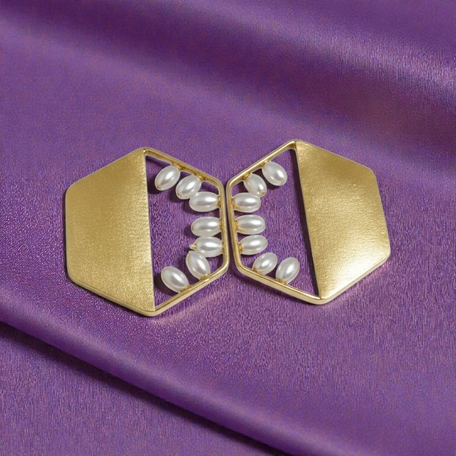 A pair of gold geometric earrings with white faux pearl accents, displayed on a purple satin fabric background.