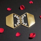 A pair of gold geometric statement earrings with white faux-pearl accents, displayed on a dark background with scattered red rose petals.
