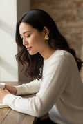 A woman with long dark hair sits at a wooden table, wearing a white knit sweater and a gold geometric stud earring. She is looking down, with a white mug partially visible in her hand, against a background of a light wall and a rustic brick wall.