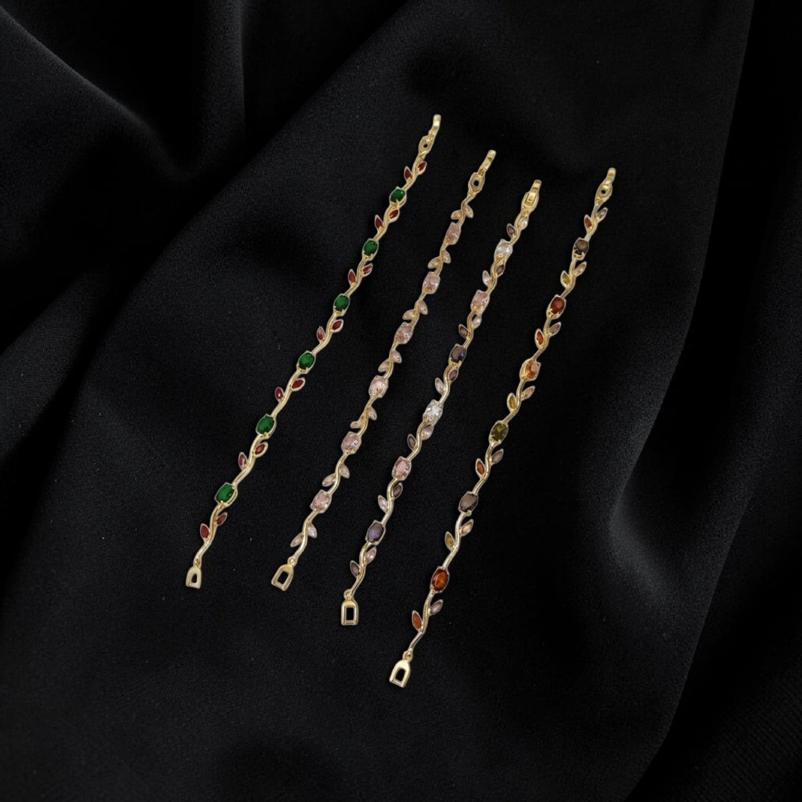 Four delicate gold-toned chain strands with small multicolored gemstone accents, arranged diagonally on a draped black fabric background.
Grishita Jewellery