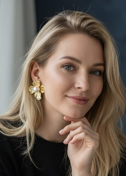 A close-up portrait of a blonde woman wearing a large, gold flower-shaped earring with a pearl center, set against a dark blue-gray background.
