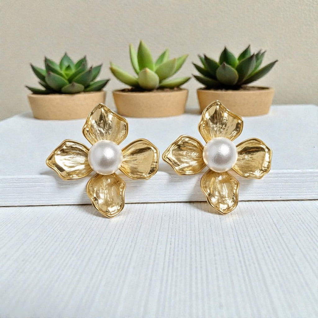 A pair of gold flower-shaped stud earrings with textured petals and a white faux-pearl center, displayed on a white wooden surface with small potted succulents in the background.