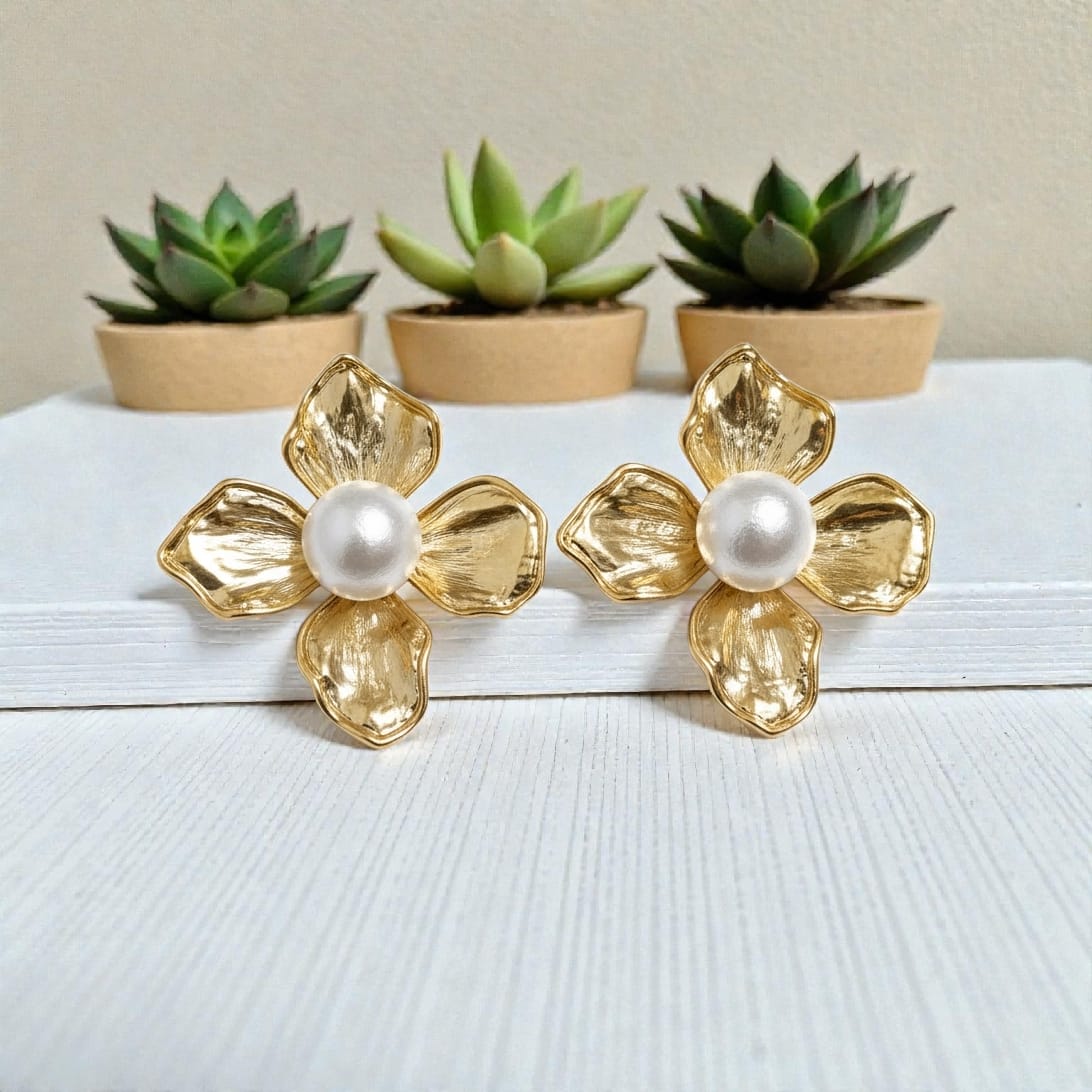 A pair of gold flower-shaped stud earrings with textured petals and a white faux-pearl center, displayed on a white wooden surface with small potted succulents in the background.