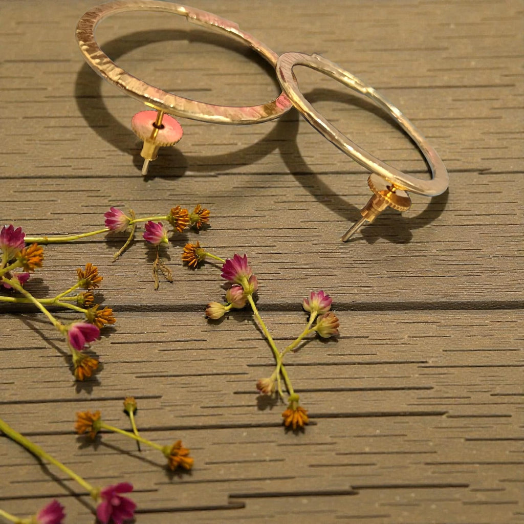 Two large textured gold hoop earrings with small pink flower-shaped dangling charms, displayed on a brown wooden surface with scattered dried flowers.