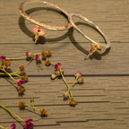 Two large textured gold hoop earrings with small pink flower-shaped dangling charms, displayed on a brown wooden surface with scattered dried flowers.