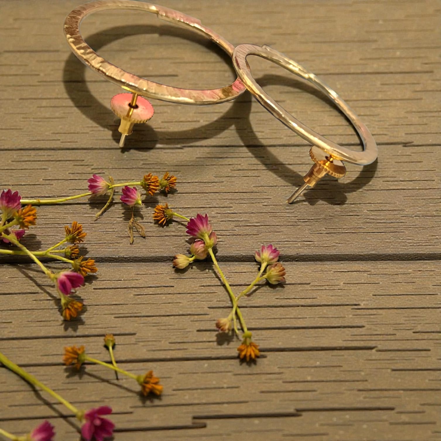 Two large textured gold hoop earrings with small pink flower-shaped dangling charms, displayed on a brown wooden surface with scattered dried flowers.