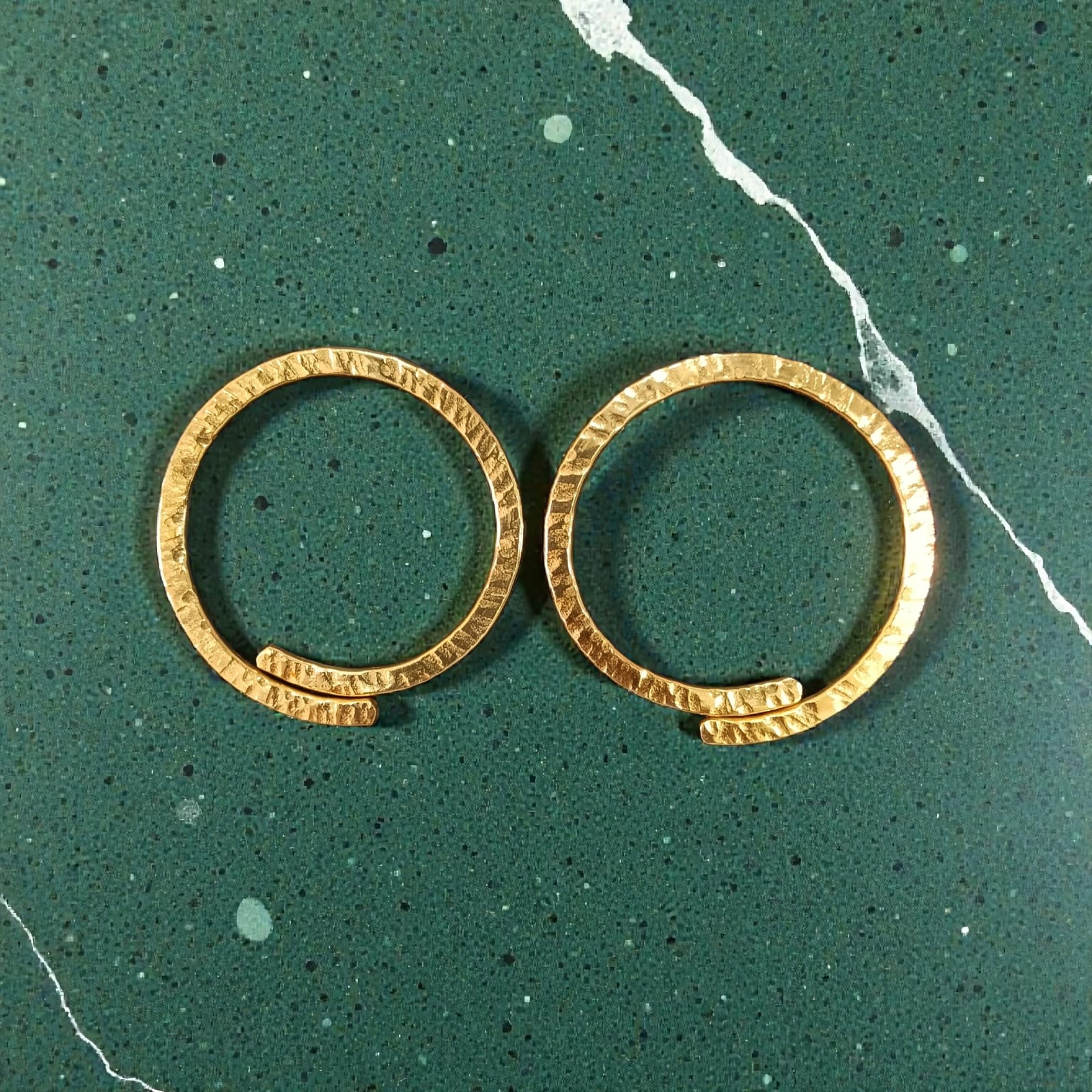 A pair of gold-toned textured hoop earrings displayed on a dark green speckled marble surface.