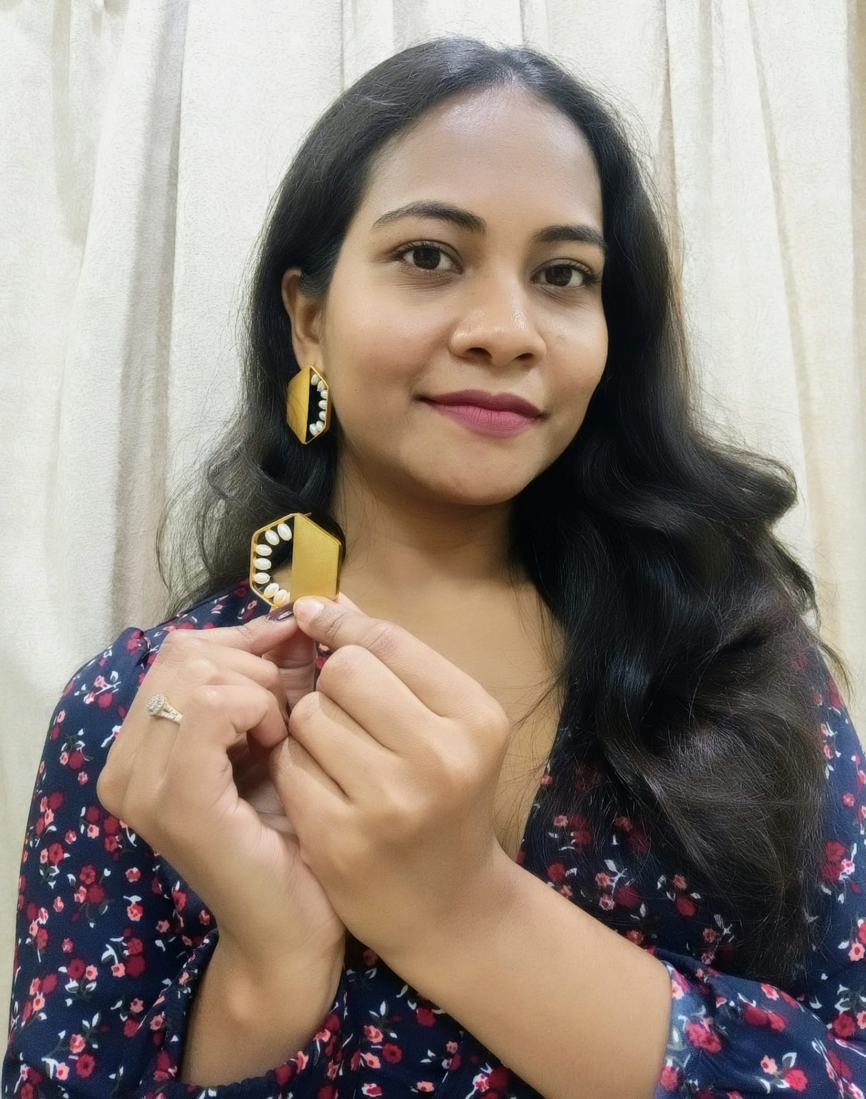 A woman with long dark hair holds up a gold and black patterned geometric earring, wearing matching gold and black earrings. She is wearing a navy blue floral top against a light beige curtain background.