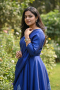 A woman wearing a royal blue long-sleeve kurta with a matching dupatta, standing in a lush green garden.