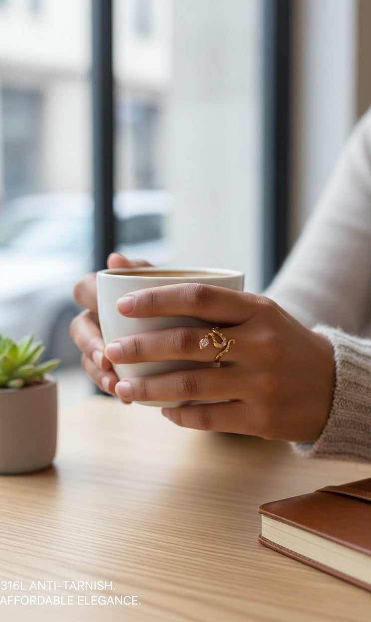 A close-up lifestyle shot of a person holding a white ceramic coffee mug on a wooden table, wearing a gold ring with a small white flower and green leaves on the ring finger.
Grishita Jewellery