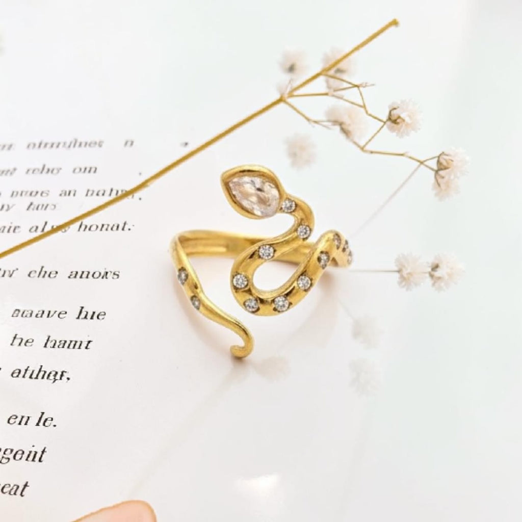 A gold snake-shaped ring decorated with clear crystals, resting on a white surface with printed text and dried baby's breath flowers.
Grishita Jewellery
