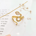 A gold snake-shaped ring decorated with clear crystals, resting on a white surface with printed text and dried baby's breath flowers.
Grishita Jewellery