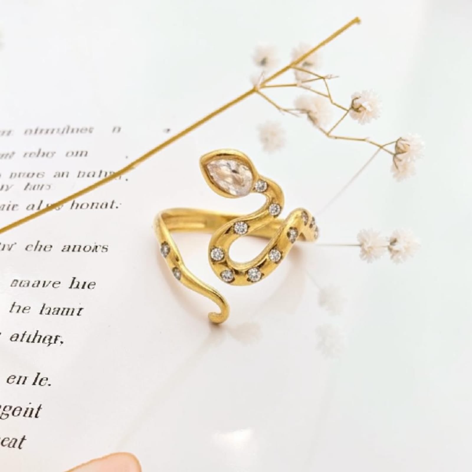 A gold snake-shaped ring decorated with clear crystals, resting on a white surface with printed text and dried baby's breath flowers.
Grishita Jewellery