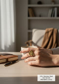A close-up lifestyle shot of a hand wearing a large, gold-toned rose-shaped ring while holding a white ceramic mug on a light wood desk. In the background, a cozy home office setting features a bookshelf, a notebook, and a pen.
Grishita Jewellery