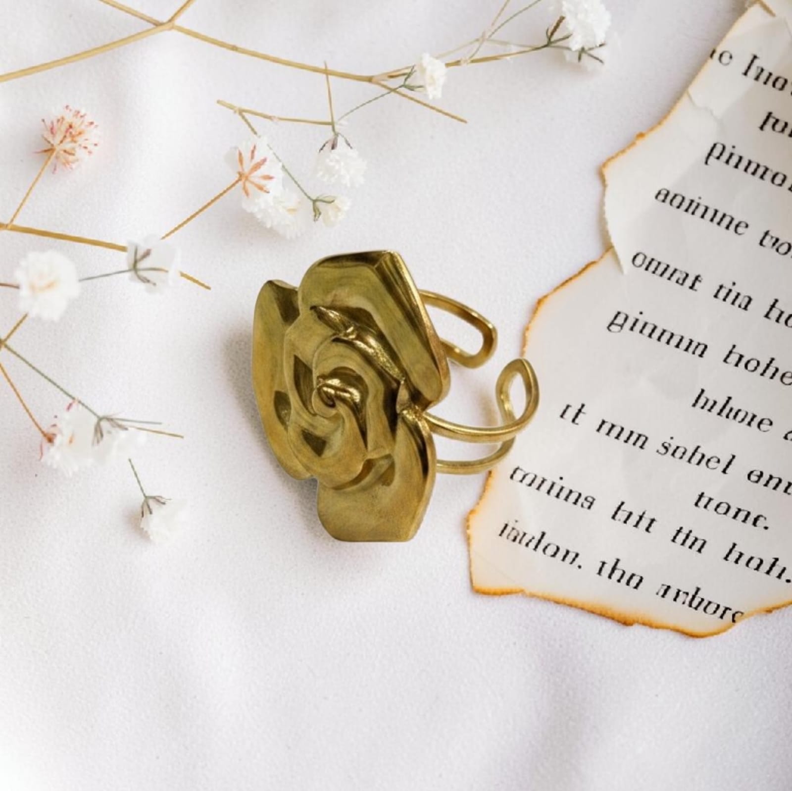 A gold-toned ring shaped like a stylized rose, photographed on a white fabric surface with small dried flowers and torn paper accents.
Grishita Jewellery