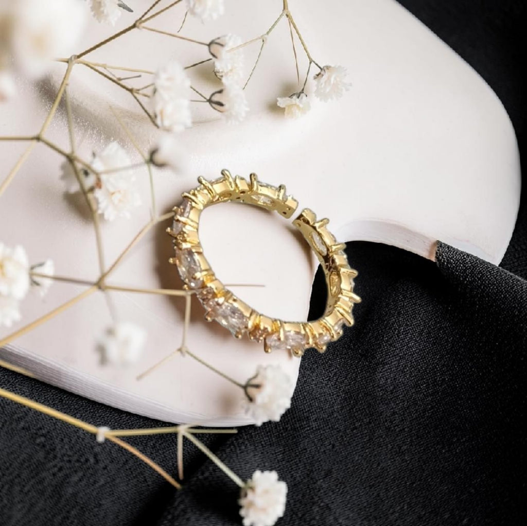 A gold ring with clear rectangular gemstones, styled on a white curved surface with white baby's breath flowers and a black fabric background.
Grishita Jewellery