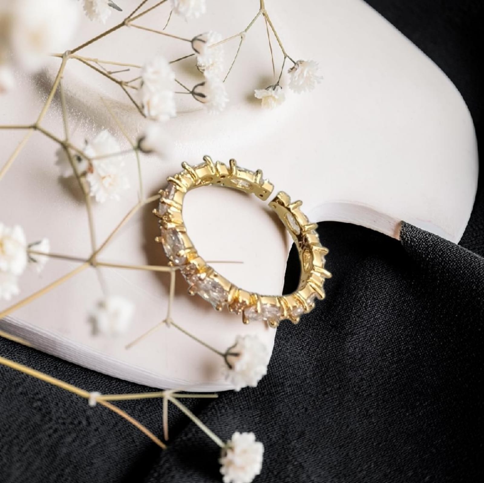 A gold ring with clear rectangular gemstones, styled on a white curved surface with white baby's breath flowers and a black fabric background.
Grishita Jewellery