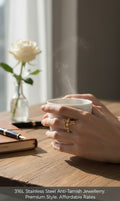 A close-up lifestyle shot of a person's hands holding a steaming white ceramic mug on a wooden table. The person is wearing a gold ring with a floral design.
Grishita Jewellery