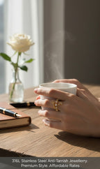 A close-up lifestyle shot of a person's hands holding a steaming white ceramic mug on a wooden table. The person is wearing a gold ring with a floral design.
Grishita Jewellery