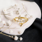 A gold ring with a butterfly motif and small clear crystal accents, styled on a white ceramic plate with dried white flowers against a black fabric background.
Grishita Jewellery