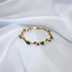 A gold leaf-patterned bracelet with green and red gemstone accents, displayed on a draped white fabric background.
Grishita Jewellery
