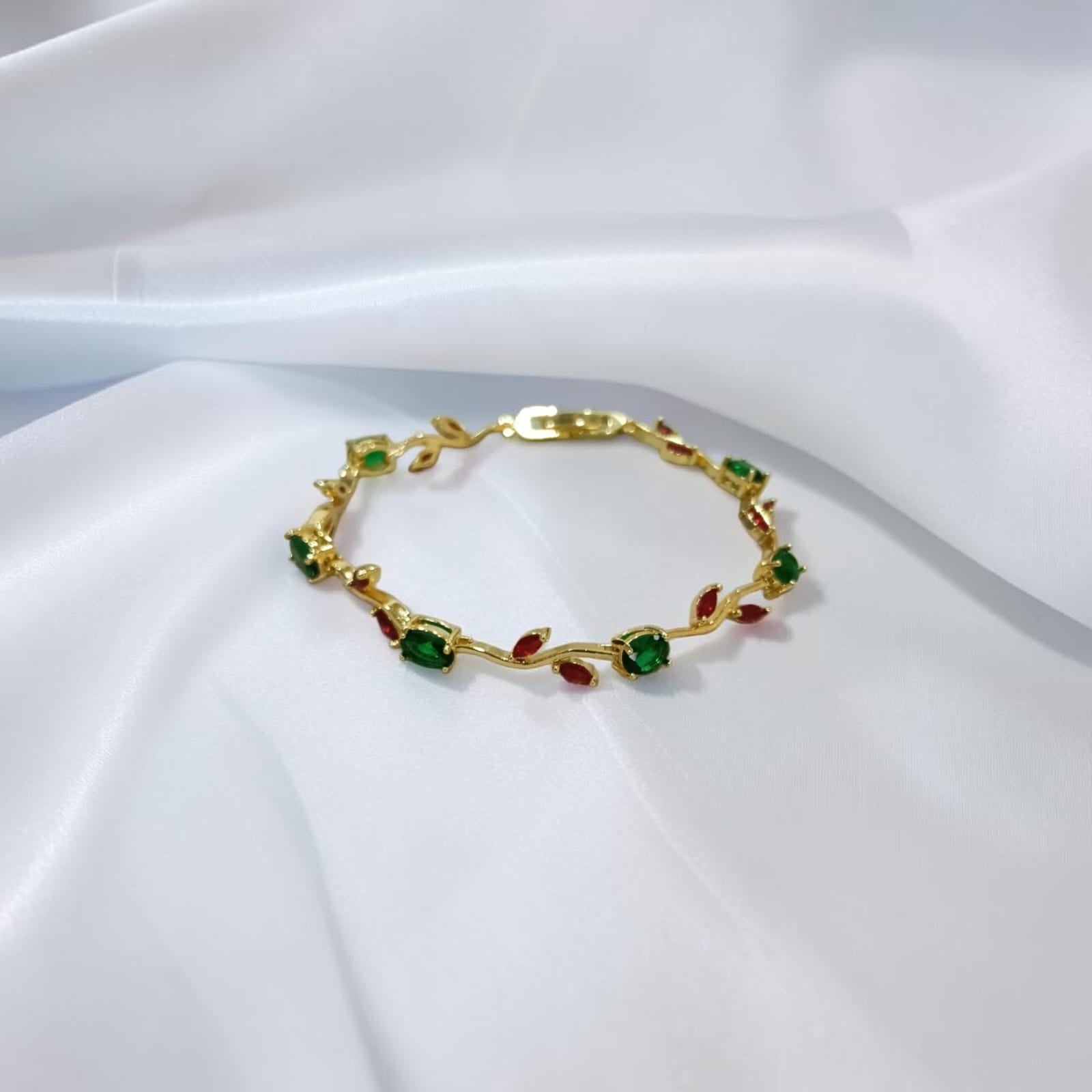 A gold leaf-patterned bracelet with green and red gemstone accents, displayed on a draped white fabric background.
Grishita Jewellery
