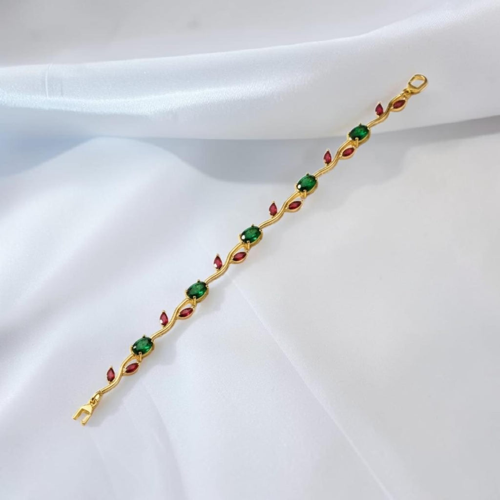 A gold-plated bracelet featuring green square gemstones and red marquise-cut gemstones, laid diagonally on a draped white satin fabric.
Grishita Jewellery