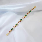 A gold-plated bracelet featuring green square gemstones and red marquise-cut gemstones, laid diagonally on a draped white satin fabric.
Grishita Jewellery