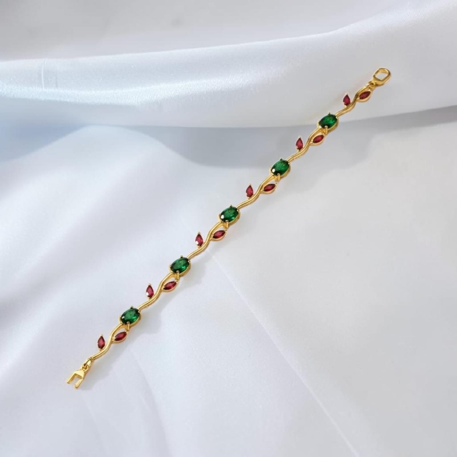 A gold-plated bracelet featuring green square gemstones and red marquise-cut gemstones, laid diagonally on a draped white satin fabric.
Grishita Jewellery