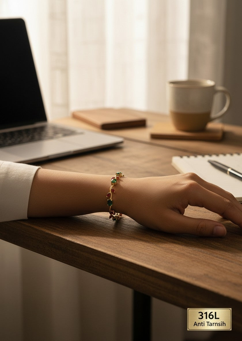 A close-up of a person's hand resting on a wooden desk, wearing a delicate gold bracelet adorned with small green and red stones. The background shows a softly lit home office environment with a laptop, notebook, and a ceramic mug.
Grishita Jewellery