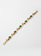 A gold-plated tennis bracelet featuring alternating green square gemstones and small red marquise-cut gemstones, displayed diagonally on a light gray background.
Grishita Jewellery