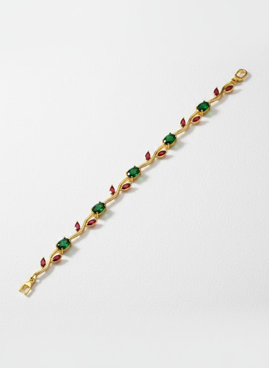 A gold-plated tennis bracelet featuring alternating green square gemstones and small red marquise-cut gemstones, displayed diagonally on a light gray background.
Grishita Jewellery