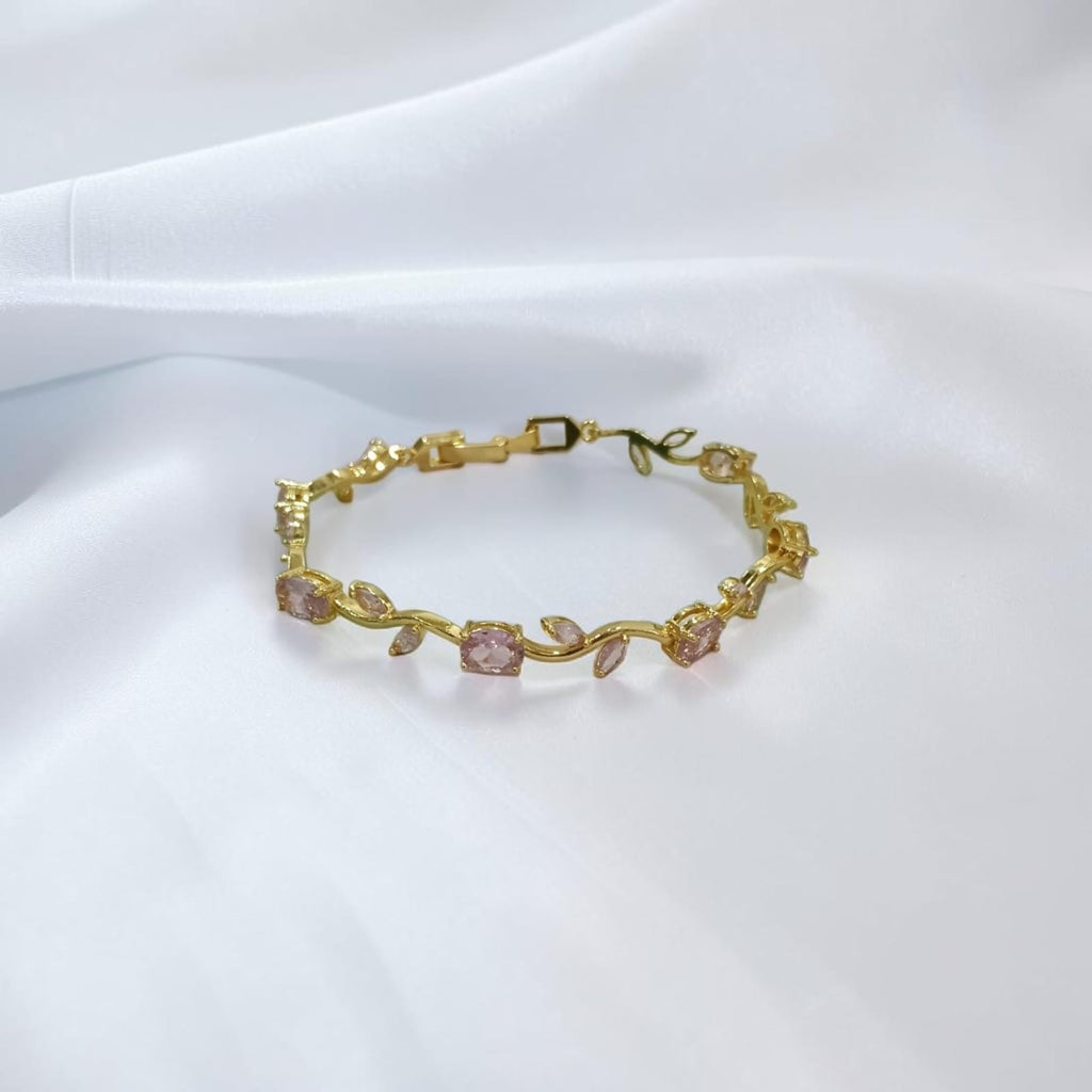 A delicate gold bracelet featuring small pink gemstone accents and leaf-shaped details, displayed on a soft white fabric background.
Grishita Jewellery