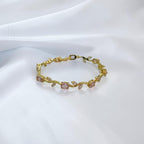 A delicate gold bracelet featuring small pink gemstone accents and leaf-shaped details, displayed on a soft white fabric background.
Grishita Jewellery