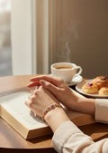 A close-up lifestyle shot of a person's hands resting on an open book, wearing a delicate gold chain bracelet with colorful gemstones. In the background, a steaming cup of coffee and pastries sit on a wooden table near a window.
Grishita Jewellery