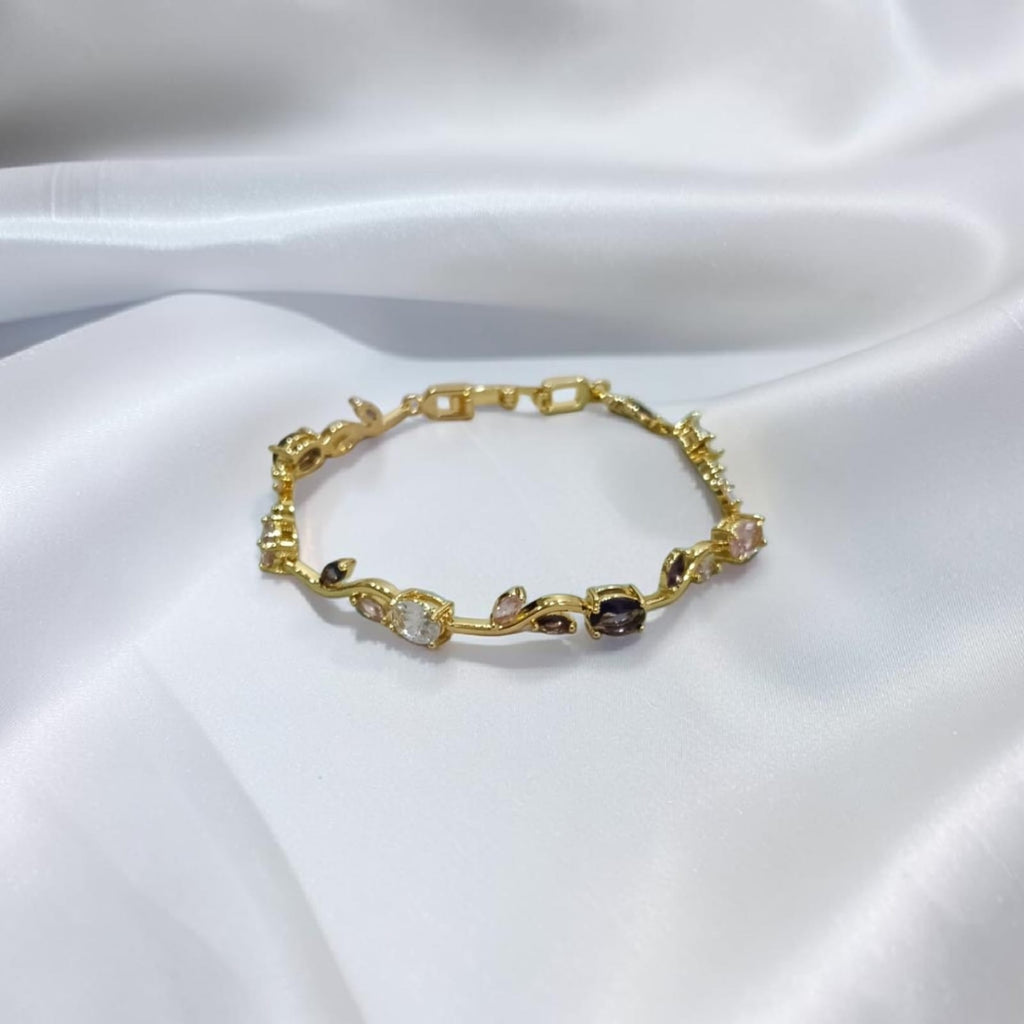 A delicate gold bracelet with small floral and geometric motifs, accented by clear and dark gemstones, displayed on a draped white satin fabric.
Grishita Jewellery