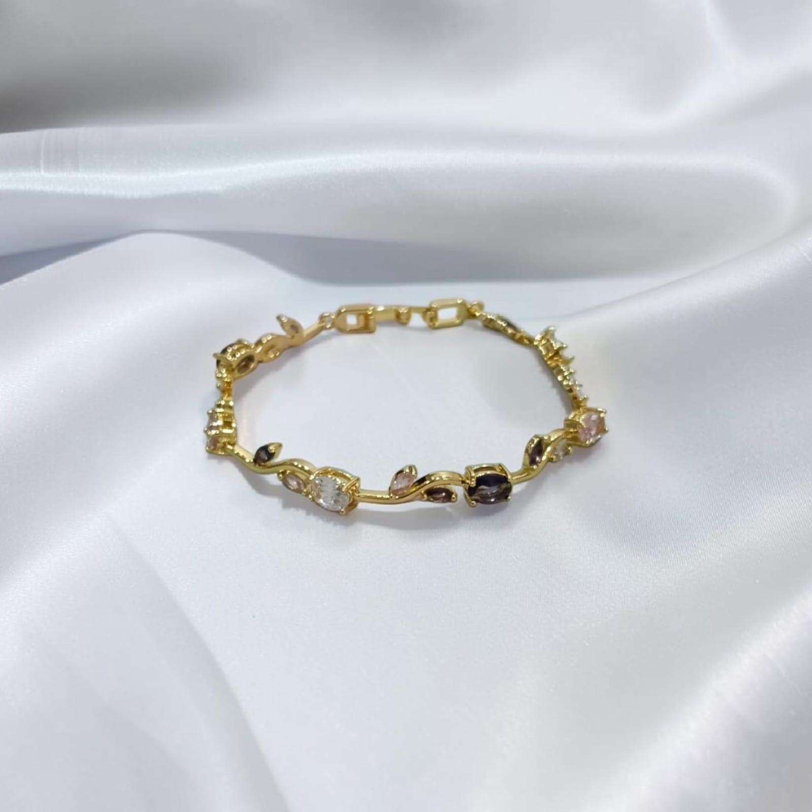 A delicate gold bracelet with small floral and geometric motifs, accented by clear and dark gemstones, displayed on a draped white satin fabric.
Grishita Jewellery