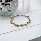 A delicate gold bracelet with leaf and vine motifs, featuring small green and peach-colored stones, displayed on a white surface with a decorative mirror ball in the background.
Grishita Jewellery