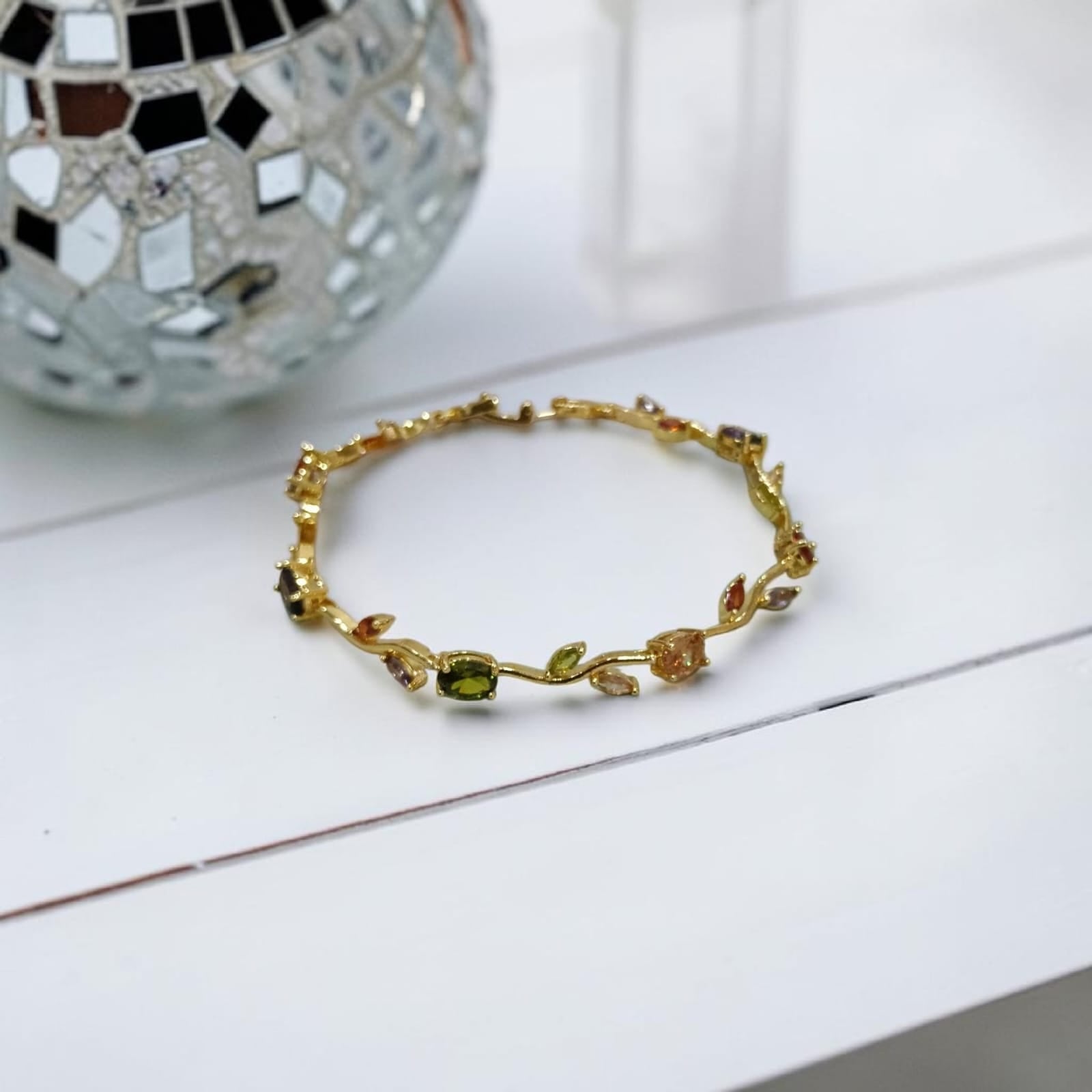 A delicate gold bracelet with leaf and vine motifs, featuring small green and peach-colored stones, displayed on a white surface with a decorative mirror ball in the background.
Grishita Jewellery