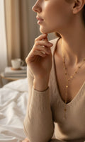A woman in a beige knit sweater wearing a delicate gold Y-necklace with small heart-shaped charms, posed in a softly lit bedroom setting.
Grishita Jewellery