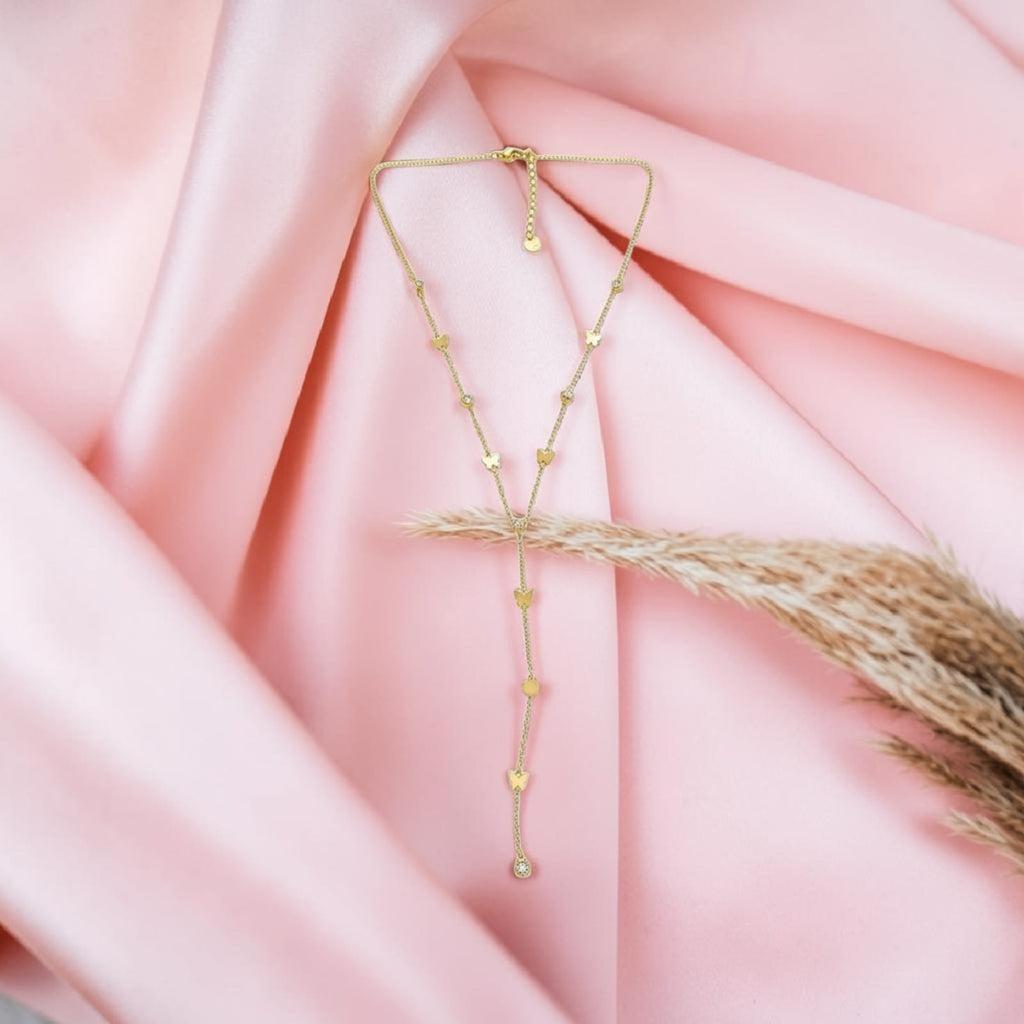 A delicate gold Y-shaped lariat necklace with small gold heart charms and a dangling heart-accented chain, displayed on draped pink satin fabric with a dried grass stem prop.
Grishita Jewellery