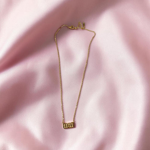 A delicate gold chain necklace with a small rectangular gold pendant engraved with the number '1111', displayed on a soft, draped pink satin fabric background.
Grishita Jewellery