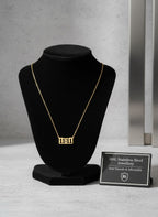 A gold rectangular pendant necklace displaying the Hebrew lettering '1111' is shown on a black velvet jewelry bust. A small black sign next to the bust reads '316L Stainless Steel Jewellery Anti-Tarnish & Affordable' with a '316L' shield logo.
Grishita Jewellery