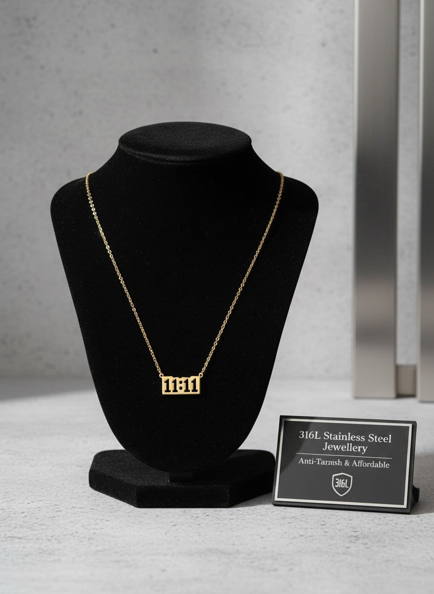 A gold rectangular pendant necklace displaying the Hebrew lettering '1111' is shown on a black velvet jewelry bust. A small black sign next to the bust reads '316L Stainless Steel Jewellery Anti-Tarnish & Affordable' with a '316L' shield logo.
Grishita Jewellery