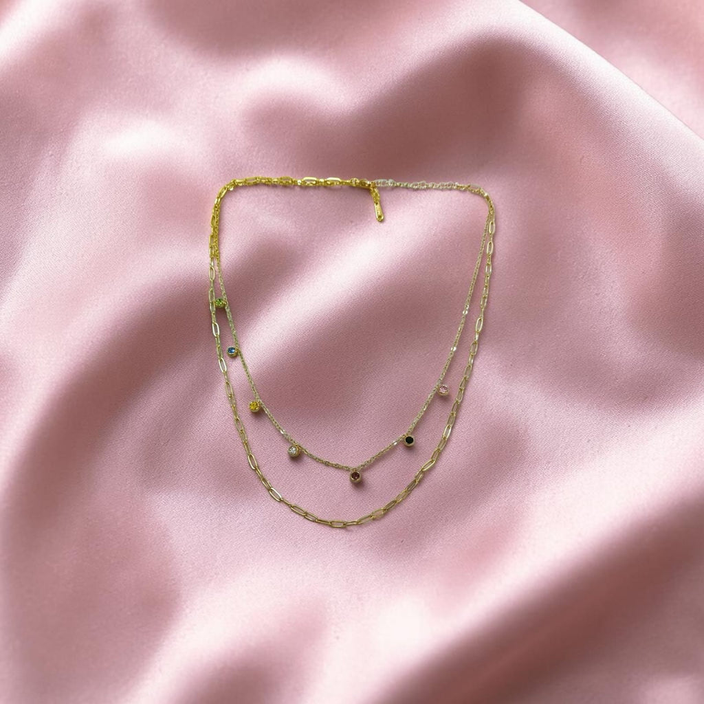 A delicate double-strand gold necklace with small green and black bead accents, laid flat on a soft pink satin fabric background.
Grishita Jewellery