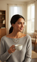 A woman with dark hair tied back wears a light gray knit sweater and layered gold necklaces, holding a white ceramic cup in a softly lit living room.