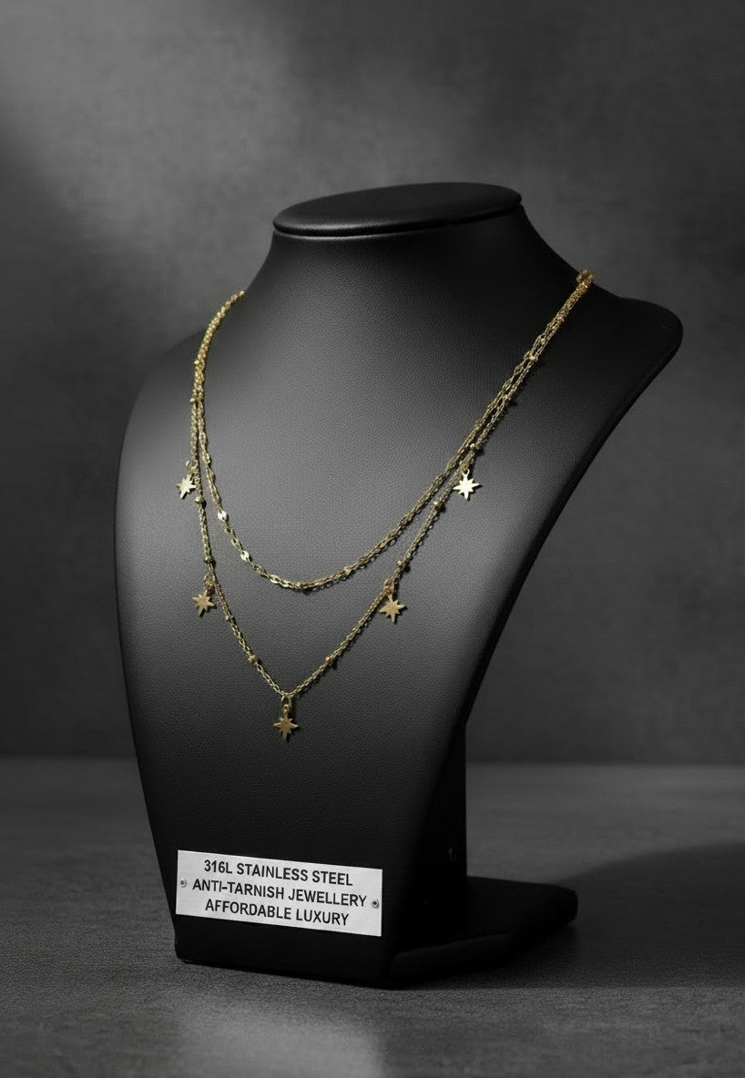 A double-layered gold necklace with small star charms displayed on a black jewelry bust. The necklace is shown against a dark gray studio background, with a small white label at the base of the bust reading '316L STAINLESS STEEL ANTI-FRAGILE JEWELLERY AFFORDABLE LUXURY'.